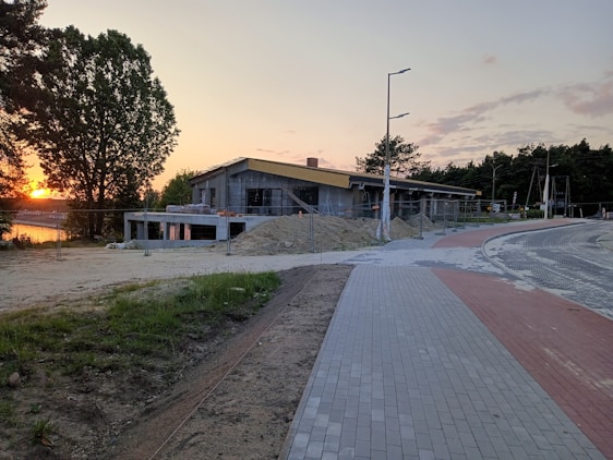 A construction site with a building being erected near a paved road, surrounded by temporary fencing. The scene is set during sunset, evident from the golden glow in the sky. Trees and shrubbery appear in the background, with a notable large tree on the left side. There are mounds of earth and building materials around the site, and the road curves away into the distance.