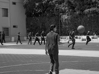 A group of people are playing basketball on an outdoor court. Several players can be seen actively dribbling and moving with the basketball. Trees and a building are in the background, with several windows visible on the building wall.