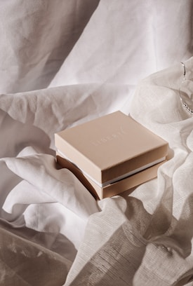 A beige box with the word 'Liberté' on top sits on a background of soft, flowing white fabric. The fabric creates a gentle, textured backdrop, suggesting an elegant and delicate setting.