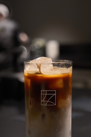 A tall glass of iced coffee with cream swirling through, topped with ice cubes. The focus is on the drink, with a dark and blurred background, giving a sense of calm and sophistication.