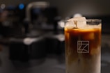A glass filled with an iced coffee beverage, topped with large ice cubes. The coffee is layered, with a darker section at the top and a lighter, milky base at the bottom. The glass features a simple geometric logo.