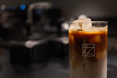 A glass filled with an iced coffee beverage, topped with large ice cubes. The coffee is layered, with a darker section at the top and a lighter, milky base at the bottom. The glass features a simple geometric logo.