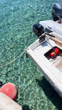 A small motorboat is moored over crystal clear water, revealing details of the rocky bottom beneath. The boat features an outboard engine labeled with brand names and a red fuel container on its deck. A red buoy floats nearby, connected by a rope.