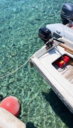A small motorboat is moored over crystal clear water, revealing details of the rocky bottom beneath. The boat features an outboard engine labeled with brand names and a red fuel container on its deck. A red buoy floats nearby, connected by a rope.