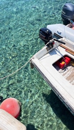 A small motorboat is moored over crystal clear water, revealing details of the rocky bottom beneath. The boat features an outboard engine labeled with brand names and a red fuel container on its deck. A red buoy floats nearby, connected by a rope.