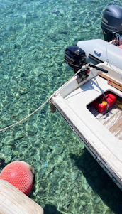A small motorboat is moored over crystal clear water, revealing details of the rocky bottom beneath. The boat features an outboard engine labeled with brand names and a red fuel container on its deck. A red buoy floats nearby, connected by a rope.