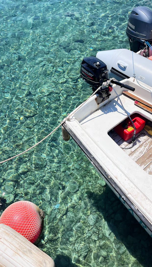 A small motorboat is moored over crystal clear water, revealing details of the rocky bottom beneath. The boat features an outboard engine labeled with brand names and a red fuel container on its deck. A red buoy floats nearby, connected by a rope.