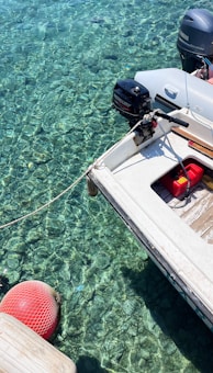 A small motorboat is moored over crystal clear water, revealing details of the rocky bottom beneath. The boat features an outboard engine labeled with brand names and a red fuel container on its deck. A red buoy floats nearby, connected by a rope.