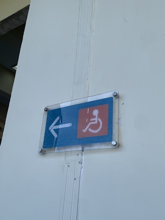 A sign mounted on a wall, displaying an arrow pointing left and a wheelchair symbol, indicating accessibility access. The sign is encased in a clear cover with visible bolts at each corner and placed against a light-colored wall.