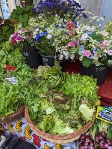 A variety of fresh greens, including lettuce and other leafy vegetables, are arranged in wicker baskets. Behind them, vibrant flowers in hues of blue, white, and pink are displayed in black containers. Price tags are visible on some of the items, suggesting a market setting.