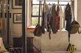 A clothing rack filled with various garments is positioned in front of a window, allowing natural light to illuminate the scene. The room has a minimalist vibe, with exposed brick walls and framed pictures. To the left, there are metal poles, possibly for photography equipment, and a radiator beneath the window. A couple of bags hang on either side of the clothing rack.