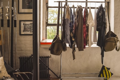 A clothing rack filled with various garments is positioned in front of a window, allowing natural light to illuminate the scene. The room has a minimalist vibe, with exposed brick walls and framed pictures. To the left, there are metal poles, possibly for photography equipment, and a radiator beneath the window. A couple of bags hang on either side of the clothing rack.