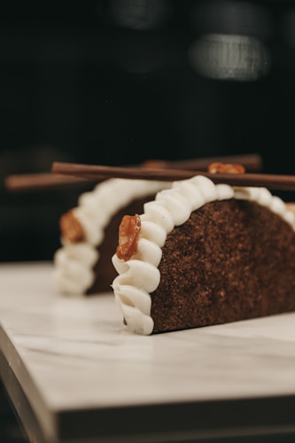 Two dessert tacos featured prominently, made with a dark, textured shell resembling chocolate or coffee flavor. Each taco is filled with a white creamy filling and garnished with caramel or brown sugar strands. Two chocolate sticks are placed on top of each taco.