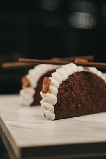 Two dessert tacos featured prominently, made with a dark, textured shell resembling chocolate or coffee flavor. Each taco is filled with a white creamy filling and garnished with caramel or brown sugar strands. Two chocolate sticks are placed on top of each taco.