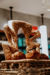 A basket filled with artisan bread, featuring a large, uniquely shaped loaf with an abstract design placed prominently. The background includes softly blurred interior lights and shelves with various items, creating a cozy atmosphere.