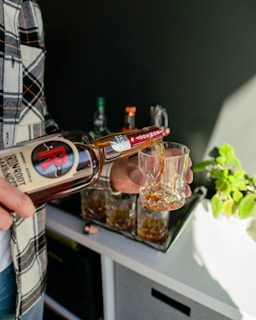 A hand pouring whisky from an elegant bottle into a glass over a rustic bar counter.