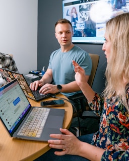 Two people are sitting at a round table, engaged in a discussion. Each has a laptop in front of them, and one person gestures while speaking. A large screen in the background displays a presentation or website.