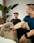 Two individuals are sitting on a beige sofa, engaged in conversation. One person is wearing a black t-shirt and cap, smiling, and has a laptop with colorful stickers. The second person, slightly blurred, is holding a mobile phone, wearing a blue t-shirt