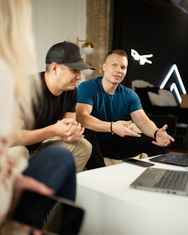 A group of individuals gathered, with one person gesturing while speaking. They are sitting around a table with laptops and smartphones. The background features a brick wall and some modern decorations, including neon lights.