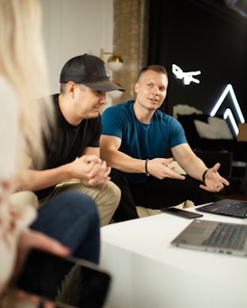 A group of individuals gathered, with one person gesturing while speaking. They are sitting around a table with laptops and smartphones. The background features a brick wall and some modern decorations, including neon lights.