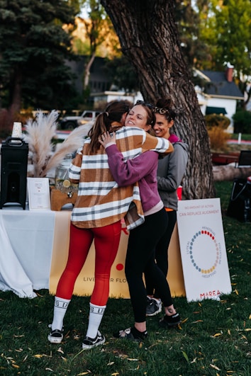 Three people are gathered outdoors, with two of them embracing warmly, suggesting a moment of friendship or reunion. They are beside a table with drinks and a tall display featuring motivational text and symbols. Green grass and trees surround the scene, indicating a park or garden setting.