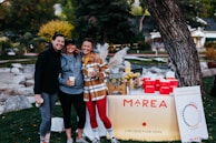 Three people are smiling and posing together outdoors in a park. They are dressed in casual, sporty clothing and holding cups. In front of them is a table with a sign that reads 'MAREA' and displays various products. The table is decorated with flowers, a glass dispenser filled with a lemon beverage, and red bags.