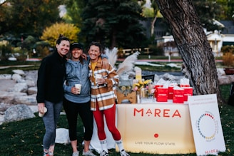 Three people are smiling and posing together outdoors in a park. They are dressed in casual, sporty clothing and holding cups. In front of them is a table with a sign that reads 'MAREA' and displays various products. The table is decorated with flowers, a glass dispenser filled with a lemon beverage, and red bags.