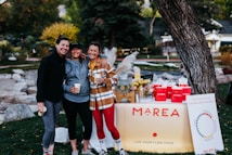 Three people are smiling and posing together outdoors in a park. They are dressed in casual, sporty clothing and holding cups. In front of them is a table with a sign that reads 'MAREA' and displays various products. The table is decorated with flowers, a glass dispenser filled with a lemon beverage, and red bags.