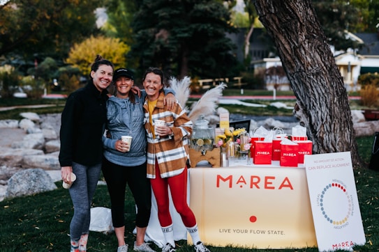 Three people are smiling and posing together outdoors in a park. They are dressed in casual, sporty clothing and holding cups. In front of them is a table with a sign that reads 'MAREA' and displays various products. The table is decorated with flowers, a glass dispenser filled with a lemon beverage, and red bags.