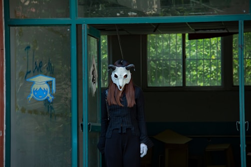 A person wearing a stylized skull mask with horns stands in a dimly lit room with windows that show greenery outside. The figure is dressed in black attire, including a long-sleeved top and matching pants. The room has a slightly eerie atmosphere, with desks and a glass door partially opened, displaying a child's drawing and the words 'I Live' written next to it.