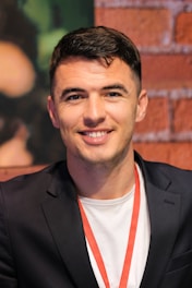 A person with short dark hair and a friendly expression is wearing a black blazer over a white shirt, with a red lanyard around the neck. The backdrop consists of a textured red brick wall and some blurred dark green elements.