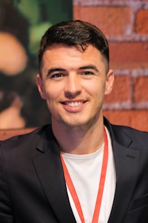 A person with short dark hair and a friendly expression is wearing a black blazer over a white shirt, with a red lanyard around the neck. The backdrop consists of a textured red brick wall and some blurred dark green elements.