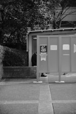 A monochrome image depicts an outdoor smoking area with a partial view of a person standing behind a panel. The panel displays signage with a smoking symbol. Surrounding the area are trees and some concrete structures.
