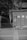 A monochrome image depicts an outdoor smoking area with a partial view of a person standing behind a panel. The panel displays signage with a smoking symbol. Surrounding the area are trees and some concrete structures.