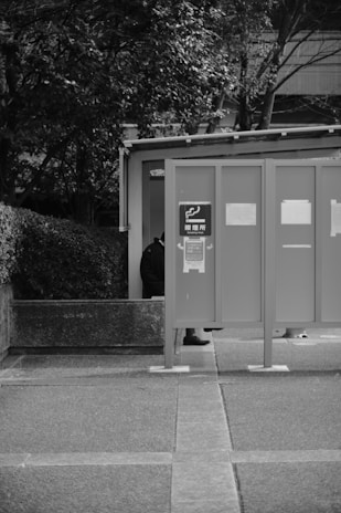 A monochrome image depicts an outdoor smoking area with a partial view of a person standing behind a panel. The panel displays signage with a smoking symbol. Surrounding the area are trees and some concrete structures.