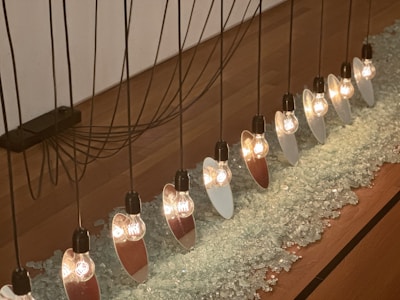 A row of light bulbs is suspended from black cables above a surface covered with crushed glass. Each bulb is accompanied by a mirrored or reflective surface, creating a unique pattern of light and reflections. The background features a wooden floor and a white wall.