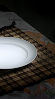 A white ceramic plate is placed on a woven bamboo placemat, which rests on a floral patterned tablecloth. The lighting creates a soft shadow around the plate, enhancing the contrast between the smooth white surface and the dark placemat.