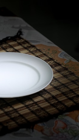 A white ceramic plate is placed on a woven bamboo placemat, which rests on a floral patterned tablecloth. The lighting creates a soft shadow around the plate, enhancing the contrast between the smooth white surface and the dark placemat.