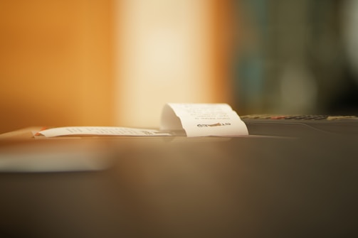 A close-up of a small paper receipt partially protruding from a machine, likely a cash register or printer. The background is softly blurred, creating a warm ambiance.