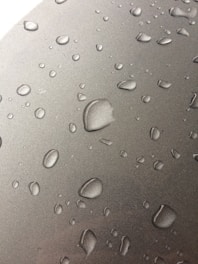 Numerous water droplets are scattered across a smooth, metallic surface. The droplets vary in size, appearing to form naturally from condensation. The surface has a reflective quality, enhancing the visibility of the droplets.