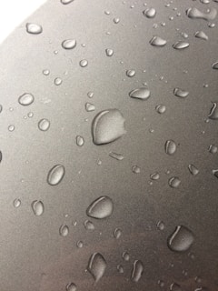 Numerous water droplets are scattered across a smooth, metallic surface. The droplets vary in size, appearing to form naturally from condensation. The surface has a reflective quality, enhancing the visibility of the droplets.