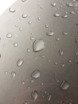 Numerous water droplets are scattered across a smooth, metallic surface. The droplets vary in size, appearing to form naturally from condensation. The surface has a reflective quality, enhancing the visibility of the droplets.