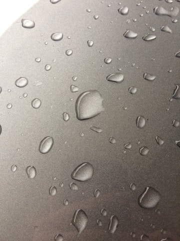 Numerous water droplets are scattered across a smooth, metallic surface. The droplets vary in size, appearing to form naturally from condensation. The surface has a reflective quality, enhancing the visibility of the droplets.