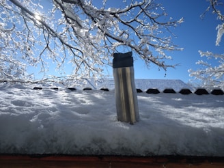 A stainless steel thermos is placed on a snowy surface with a backdrop of snow-covered branches and a clear blue sky. Sunlight is creating a bright reflection on the snow and the thermos.