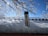A stainless steel thermos is placed on a snowy surface with a backdrop of snow-covered branches and a clear blue sky. Sunlight is creating a bright reflection on the snow and the thermos.