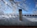 A stainless steel thermos is placed on a snowy surface with a backdrop of snow-covered branches and a clear blue sky. Sunlight is creating a bright reflection on the snow and the thermos.