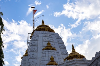 A temple with intricate white carvings and golden domes set against a bright blue sky with scattered clouds. A flag with a red background is mounted on a pole at the top of the temple.