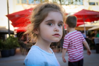 A young child with brown hair is looking towards the camera with a neutral expression. Another child, wearing a striped shirt, is in the background facing away. Red umbrellas and an outdoor seating area of a cafe or restaurant are seen, suggesting a sunny day.