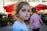 A young child with brown hair is looking towards the camera with a neutral expression. Another child, wearing a striped shirt, is in the background facing away. Red umbrellas and an outdoor seating area of a cafe or restaurant are seen, suggesting a sunny day.