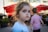 A young child with brown hair is looking towards the camera with a neutral expression. Another child, wearing a striped shirt, is in the background facing away. Red umbrellas and an outdoor seating area of a cafe or restaurant are seen, suggesting a sunny day.